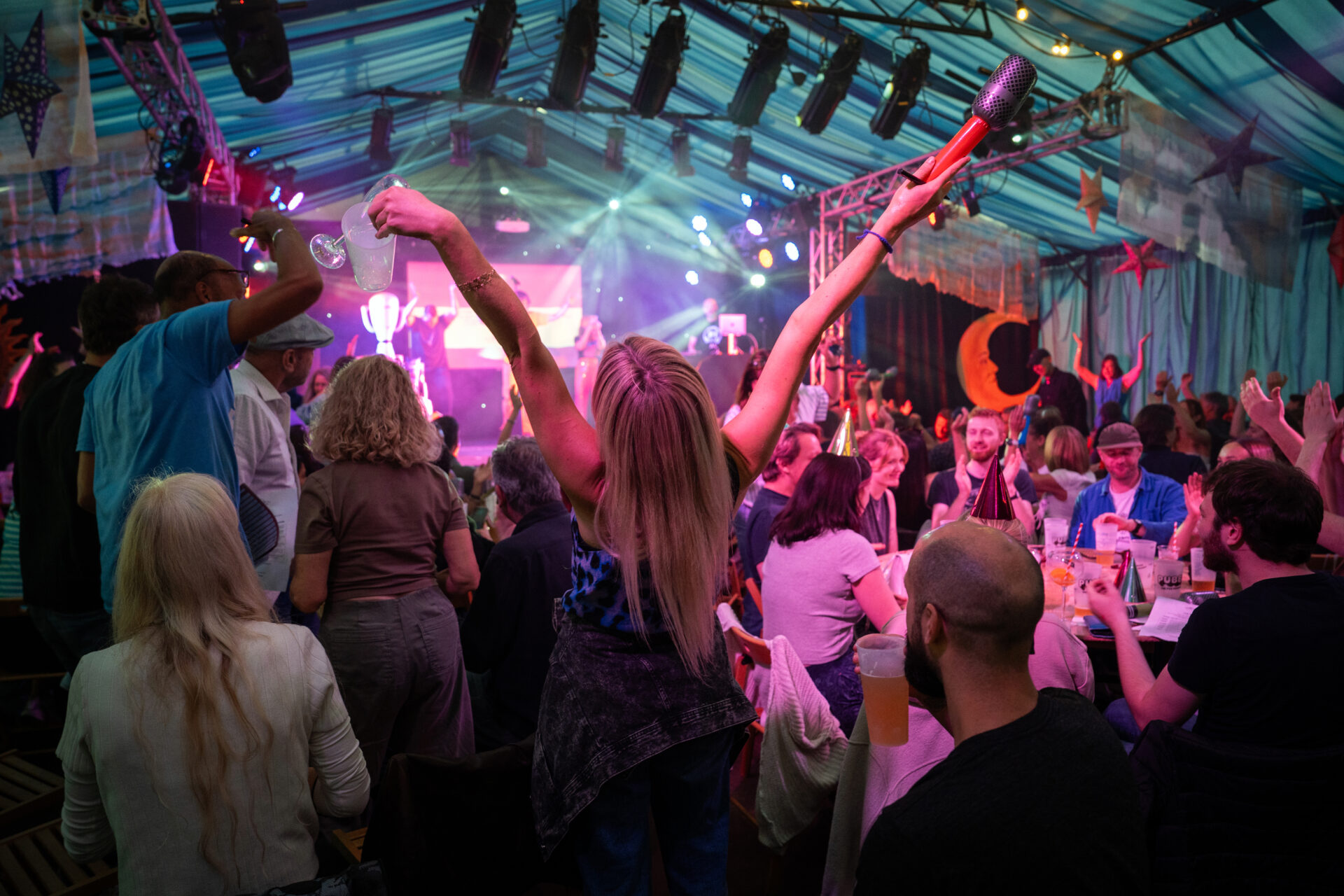 An audience member in a crowd with their arms above their head cheering at the performance in front of them